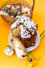 Easter cake with white and black chocolate sauce, spring flowers and an egg on a bright yellow tablecloth, behind an Easter basket