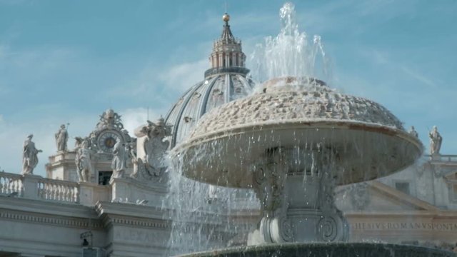 Low angle view camera tracking right past water fountain with view of the dome of St. Peter's Basilica in Vatican City
