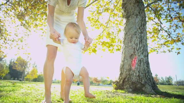 European mother supporting her little baby son as he practices his first steps outdoors on the grass 