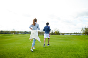 Rear view of active girl and guy in sportswear running down green grass in the morning