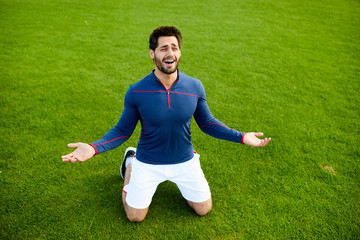 Active young man in sportswear standing on his knees on green field with his hands open