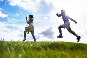 Active young couple running down green grass against cloudy sky on summer morning