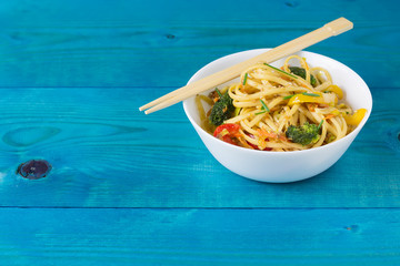 Asian food. Stir fry udon noodles with vegetables on a white bowl, blue wooden backgound, cooked in...