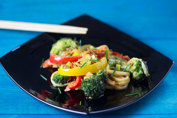 Asian food. Stir fry udon noodles with vegetables on a black plate, blue wooden backgound, cooked...