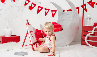 portrait of a cute little baby girl in a decorated holiday studio of the Valentine's day . Child girl with blond hair sitting and holding a brush draws a heart.