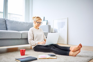 Cheerful woman sitting on floor at home having coffee and using laptop