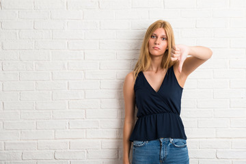 Blonde woman showing thumb down sign with negative expression on white brick wall background