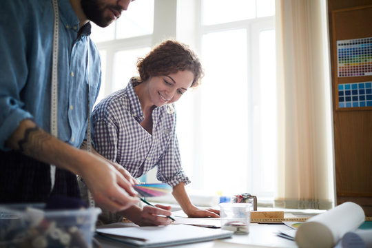 Young woman leaning over sketch and looking at color swatches held by colleague