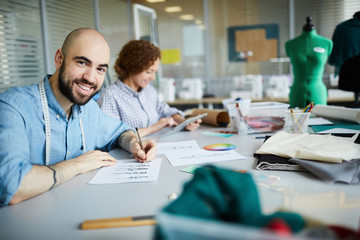 Happy young designer sitting by desk and sketching new fashion models in working environment