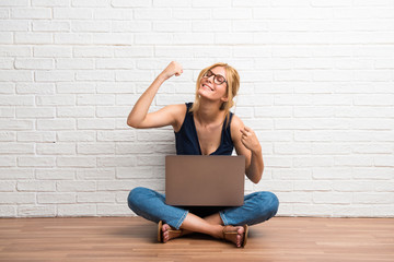 Blonde girl sitting on the floor with her laptop celebrating a victory on white brick wall background