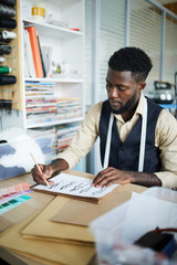 Young fashion designer drawing sketches of new models on paper in his workshop or studio