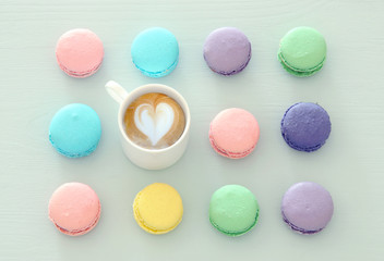 Top view image of white cup of coffee and colorful macaron or macaroon over pastel wooden background.