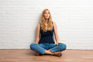 Blonde girl sitting on the floor makes funny and crazy face emotion on white brick wall background