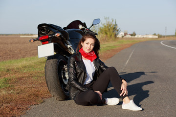 Obraz premium Relaxed carefree female motorcyclist wears black clothes and white trainers, sits at asphalt near motorbike, enjoys adventure and racing in countryside, prepares for motorcross. Leisure concept