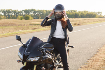 Outside shot of fast female motorcyclist wears protective helmet, poses on motorbike, stands on...