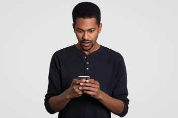 Astonished black man with curly hair, holds mobile phone, recieves text message, feels surprised and amazed, wears casual clothes, isolated over white background. People, reaction and technology