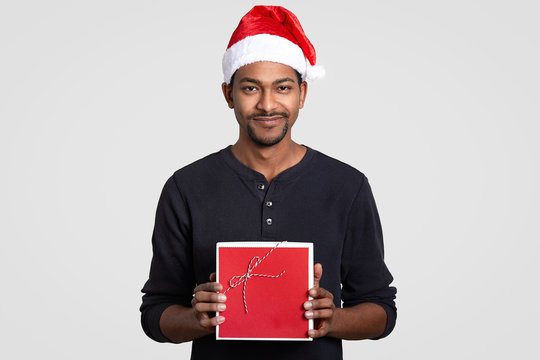 Horizontal shot of pleasant looking male holds red present, wears festive hat, recieves present on New Year or Christmas, models over white background. Santa Claus with gift box. Celebration concept