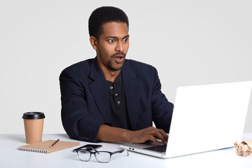 Shocked businessman with black skin, keyboards information on laptop computer, stupefied to fail make presentation, drinks coffee, writes down information in notepad, isolated over white background