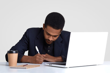 Busy male worker with dark healthy skin, writes down information in papers, holds pen, wears black suit, rewrites information from laptop computer, drinks coffee, isolated over white background