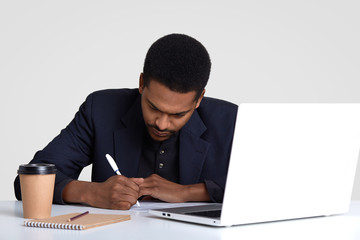 Creative copywriter writes down ideas, sits in front of opened laptop computer, surrounded with...