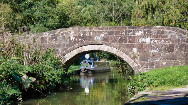 Looking at a peaceful canal scene with a boat moored and framed by an old stone bridge. The reflections are in the water