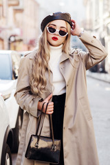 Outdoor fashion portrait of young beautiful woman wearing trendy beige trench coat, stylish cat eye sunglasses, leather beret, turtleneck, holding small bag, model posing in street of european city