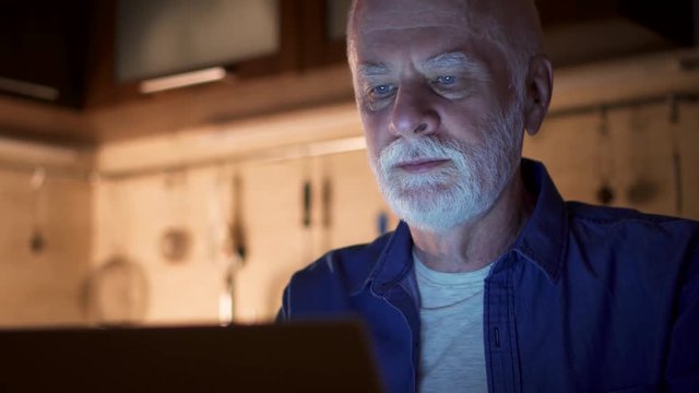 Senior man relax at home reading news on laptop. Male freelancer using computer at night working hard from home office to meet deadline. Overworked businessman working on project in kitchen in evening