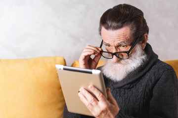 Senior bearded man in glasses wearing grey pullover sitting on a yellow sofa in his light living room and looking over his glasses and reading something on the tablet computer.