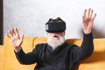 Naklejka premium Contemporary senior bearded man in grey pullover testing VR device while sitting on the yellow sofa in the light living room, future technology concept