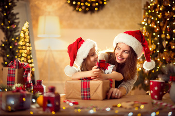 Happy mother and little girl in santa helper hat pack Christmas gifts and have fun over living room, christmas tree, lights background