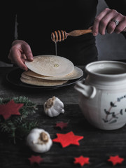 Female hands pouring honey on Christmas Eve wafers. Pot of honey and garlic bulbs on dark wooden table. 