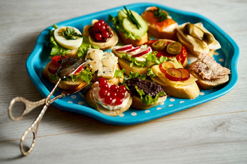 old blue tray with a set of healthy tapas or mini sandwiches with food tongs on a light wooden table