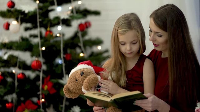 The mum is reading the book to small daughter and kissing her on cheek while girl's playing a teedy bear. Both female  dressed in cherry dresses  There is Christmas tree background.