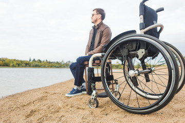 A young man sits on a bench by the lake, next to his wheelchair.