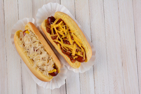 Two hot dogs one with sauerkraut and another with chili cheese toppings on a white ewood table with copy space.