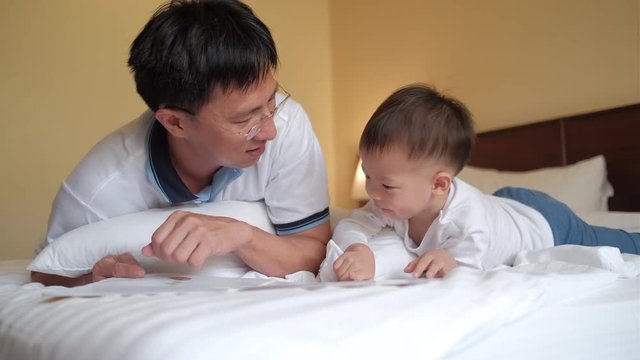 Smiling father and cute little Asian 2 years old toddler baby boy child reading bedtime story book, sitting on bed at home, Dad and son spending time together, Parents reading to child concept