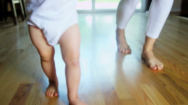 Young Caucasian mother supporting her little baby son as he practices his first steps indoor 