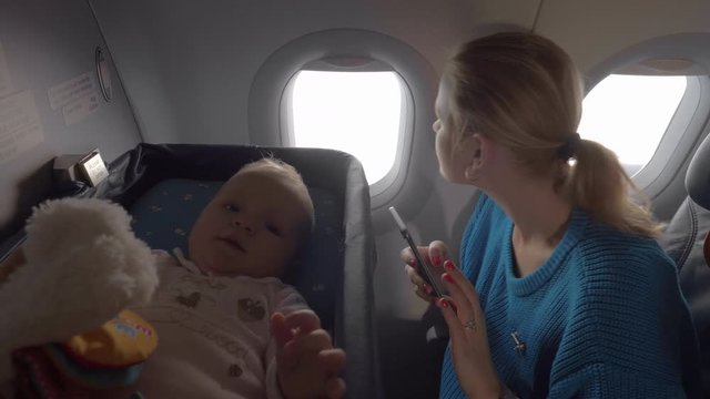 Medium shot of a cute baby in a baby cot bassinet on board an airplane while her mother checks her cellphone