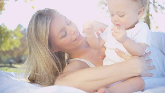 Loving beautiful blonde Caucasian mom lying outdoors on the blanket playing with her young baby son 