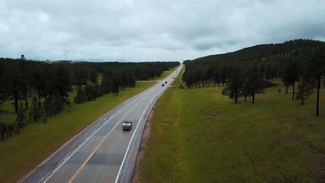 Drone flying low over traffic on highway road between wild clean green forest hills with trees on cloudy summer day.