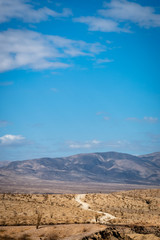Unpaved road winds through desert landscape toward mountains and cloudy blue skies.