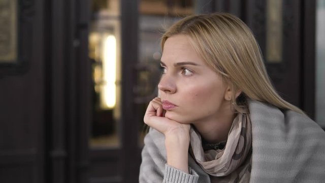 A young beautiful blonde trendy dressed woman thinking about something resting her chin on her arm sitting in a cozy street cafe.