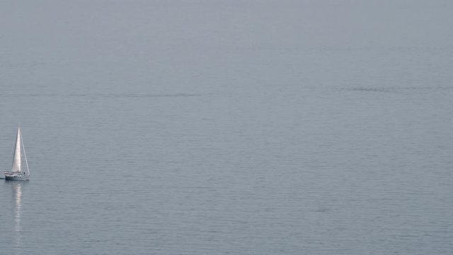 A lonely white yacht is slowly floating on the quite sea surface. The day is clear, there is no wind so nothing is bothering the water and the white sail