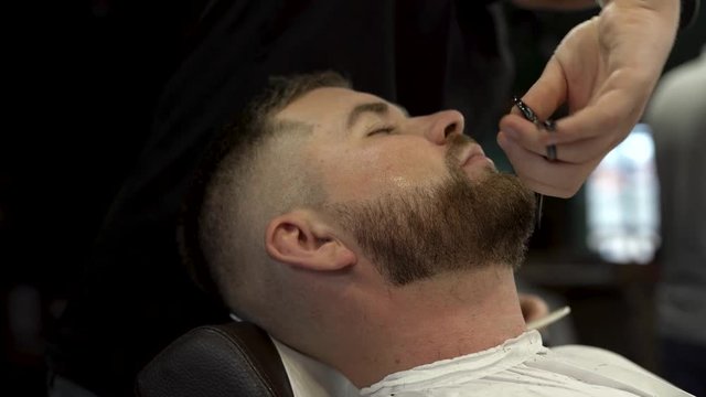 Barber tattooed hands with black torso are cutting client's brown beard using a comb and scissors. The customer is with the eyes closed and coverd with white hairdresser peignoir