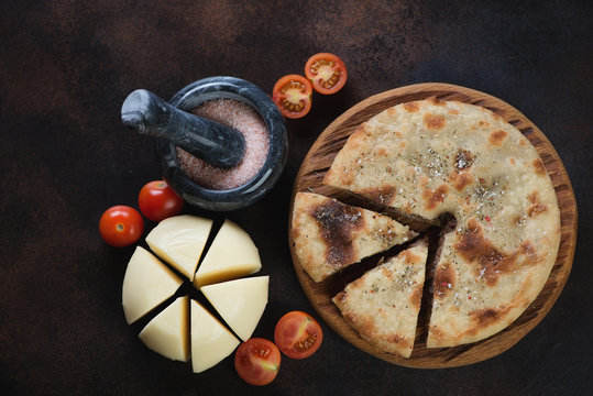 Top view of sliced ossetian pie, cherry tomatoes and suluguni on a dark brown metal background, horizontal shot