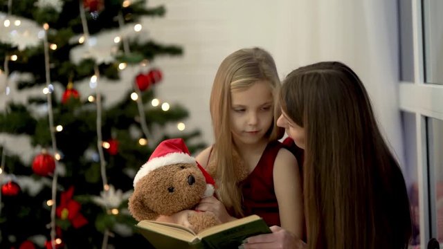 The mum is reading the book to small daughter, girl's holdingg a teedy bear and starring at mum. Both female  dressed in cherry dresses  There is Christmas tree background.