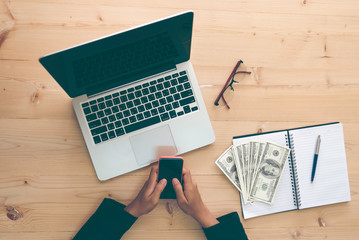  Female entrepreneur working and typing on smartphone at her office wooden desk with glasses, money and pennon notebook and laptop laptop in high angle view for work at home, yolo, technology concept