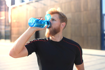 Handsome adult man drinking water from fitness bottle while standing outside, at sunset or sunrise....