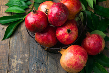 Ripe juicy organic bright red pomegranates in metal wicker basket with branches green leaves on plank barn wood background. Fall produce harvest vitamins abundance concept. Rosh Hashanah
