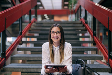 Young Caucasian female student with brown hair and eyeglasses using tablet while sitting on the stairs in college building. Next to her bag. - obrazy, fototapety, plakaty